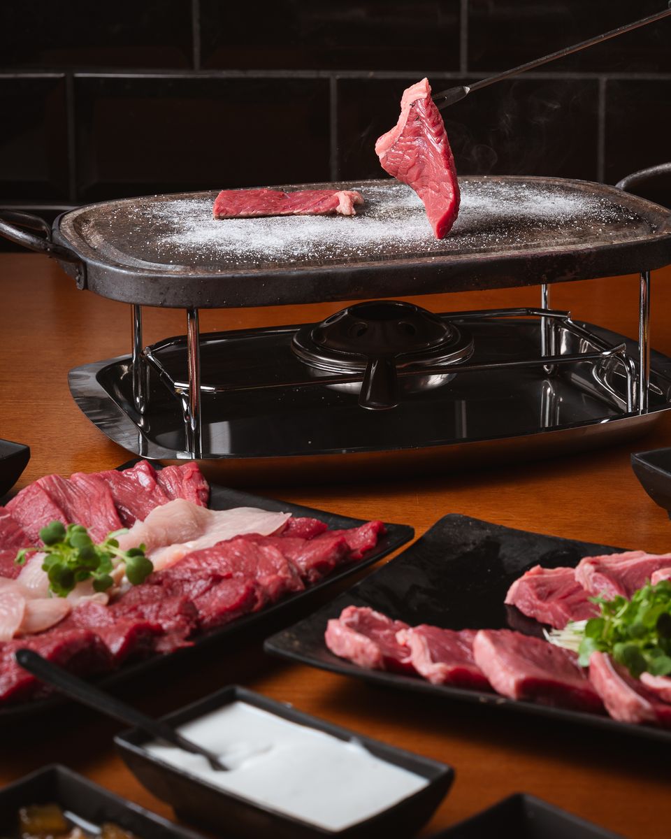 Carne sendo virada na pedra quente com sal grosso, com travessas de carnes e frango e potes de molho na mesa